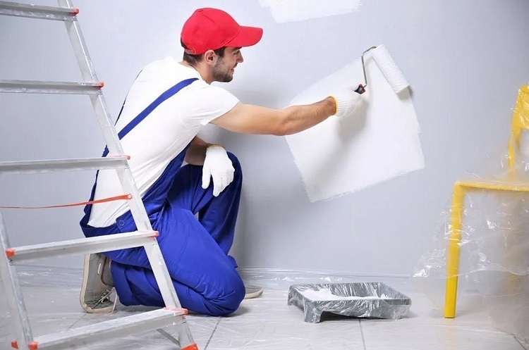 Young man wearing red cap and blue jumper holding a painting roller working in a room